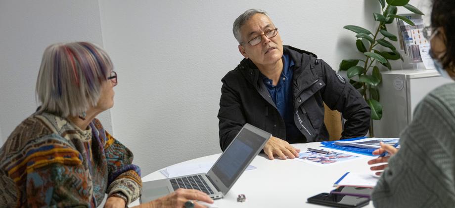Bir masada iki hanımefendi ve bir beyefendi konuşuyor. Soldaki hanımefendinin önünde açık bir bilgisayar var.