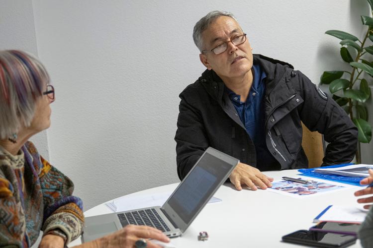 Bir masada iki hanımefendi ve bir beyefendi konuşuyor. Soldaki hanımefendinin önünde açık bir bilgisayar var.