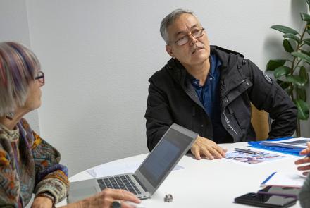 Bir masada iki hanımefendi ve bir beyefendi konuşuyor. Soldaki hanımefendinin önünde açık bir bilgisayar var.